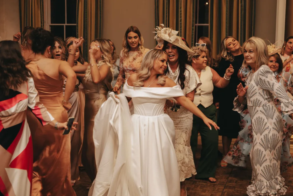 A joyful bride in an off-shoulder white gown dances surrounded by women in elegant dresses at Rudding Park. The room, with large windows and striped curtains, is a lively scene. In Harrogate's charming setting, guests in formal and floral outfits—some with elaborate hats—celebrate merrily. © Aimee Lince Photography - Wedding photographer in Lincolnshire, Yorkshire & Nottinghamshire