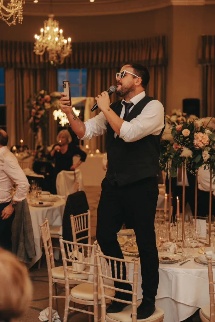 At Rudding Park in North Yorkshire, a man in a white shirt, black vest, and tie stands on a chair, holding a microphone and smartphone. Indoors with sunglasses on, he's either singing or speaking at this formal Harrogate event with elegantly set tables and chandeliers overhead. © Aimee Lince Photography - Wedding photographer in Lincolnshire, Yorkshire & Nottinghamshire