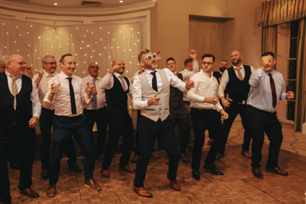 A group of men in formal attire joyfully dancing indoors at a Harrogate venue. The central figure, sporting white heart-shaped sunglasses and a vest, leads the energetic movement. Behind them, warm lighting and a decorated curtain create a lively atmosphere reminiscent of Rudding Park in North Yorkshire. © Aimee Lince Photography - Wedding photographer in Lincolnshire, Yorkshire & Nottinghamshire