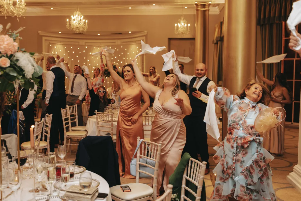 At a formal event in Harrogate, guests joyfully wave napkins in the air. Women in elegant satin and floral dresses are smiling and laughing. The setting at Rudding Park features white chairs, floral arrangements, and warm lighting with chandeliers. The atmosphere is lively and celebratory. © Aimee Lince Photography - Wedding photographer in Lincolnshire, Yorkshire & Nottinghamshire