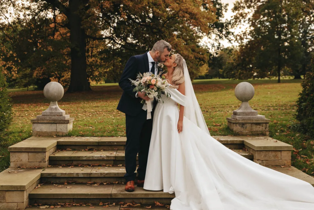 A bride and groom share a kiss on stone steps surrounded by greenery at Rudding Park. The bride, in a flowing white gown with a veil, holds a bouquet of flowers. The groom is in a navy suit. Autumn leaves are scattered on the ground in North Yorkshire, with trees under an overcast Harrogate sky. © Aimee Lince Photography - Wedding photographer in Lincolnshire, Yorkshire & Nottinghamshire
