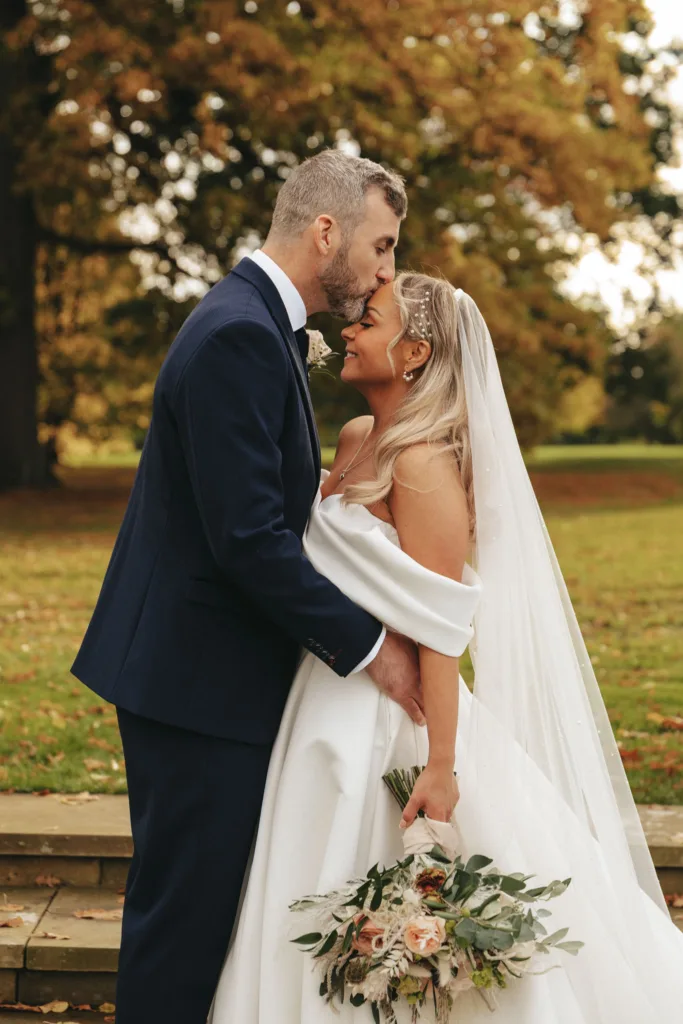 A groom in a dark suit kisses a bride's forehead as they stand outdoors in North Yorkshire. The bride, in a white off-shoulder gown, holds a bouquet of pink and white flowers and smiles up at him. They're surrounded by green grass and trees with autumn leaves at Rudding Park. © Aimee Lince Photography - Wedding photographer in Lincolnshire, Yorkshire & Nottinghamshire