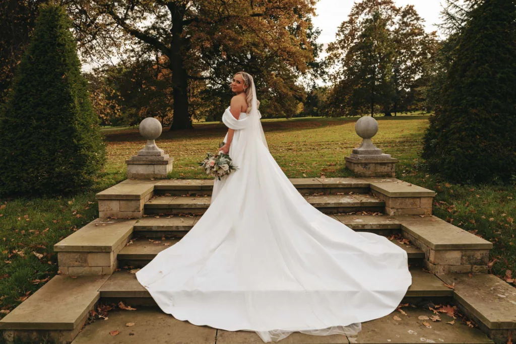 In the gardens of Rudding Park, a bride in a flowing white gown stands on stone steps, her bouquet clutched tightly. She gazes over her shoulder as her long train cascades down the steps, surrounded by green bushes and trees with autumn leaves in Harrogate, North Yorkshire. © Aimee Lince Photography - Wedding photographer in Lincolnshire, Yorkshire & Nottinghamshire