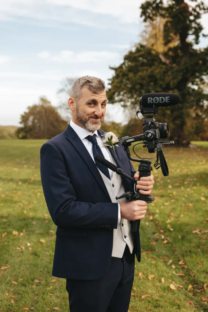 A man with graying hair and a beard is wearing a dark blue suit with a white shirt and gray vest, holding a professional camera with a "RODE" microphone. He stands on the grassy field of Rudding Park, North Yorkshire, among scattered autumn leaves and trees under a partly cloudy sky. © Aimee Lince Photography - Wedding photographer in Lincolnshire, Yorkshire & Nottinghamshire