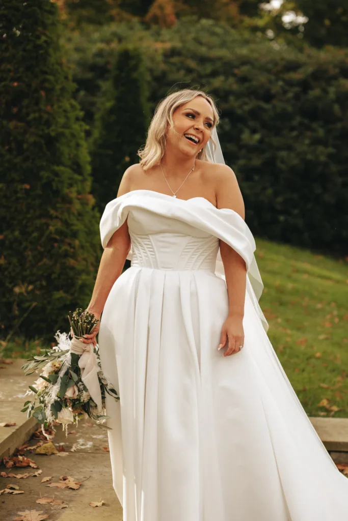 In the lush greenery of Rudding Park, a bride in an off-the-shoulder white wedding gown stands outdoors, smiling. She holds a bouquet of white flowers and greenery. Her blond hair is styled down with a veil, while the slightly blurred path in North Yorkshire adds a magical touch. © Aimee Lince Photography - Wedding photographer in Lincolnshire, Yorkshire & Nottinghamshire
