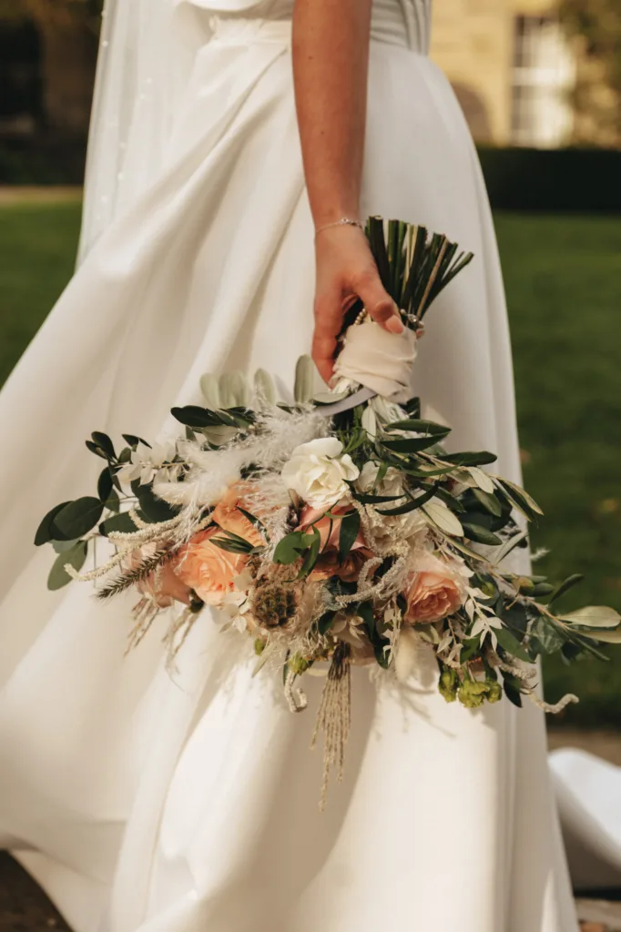 A bride in a white dress holds a bouquet, featuring peach roses and pampas grass. She stands on a grassy lawn at Rudding Park, with North Yorkshire's serene beauty as her backdrop. The blurred building and garden add to the tranquil Harrogate setting. © Aimee Lince Photography - Wedding photographer in Lincolnshire, Yorkshire & Nottinghamshire