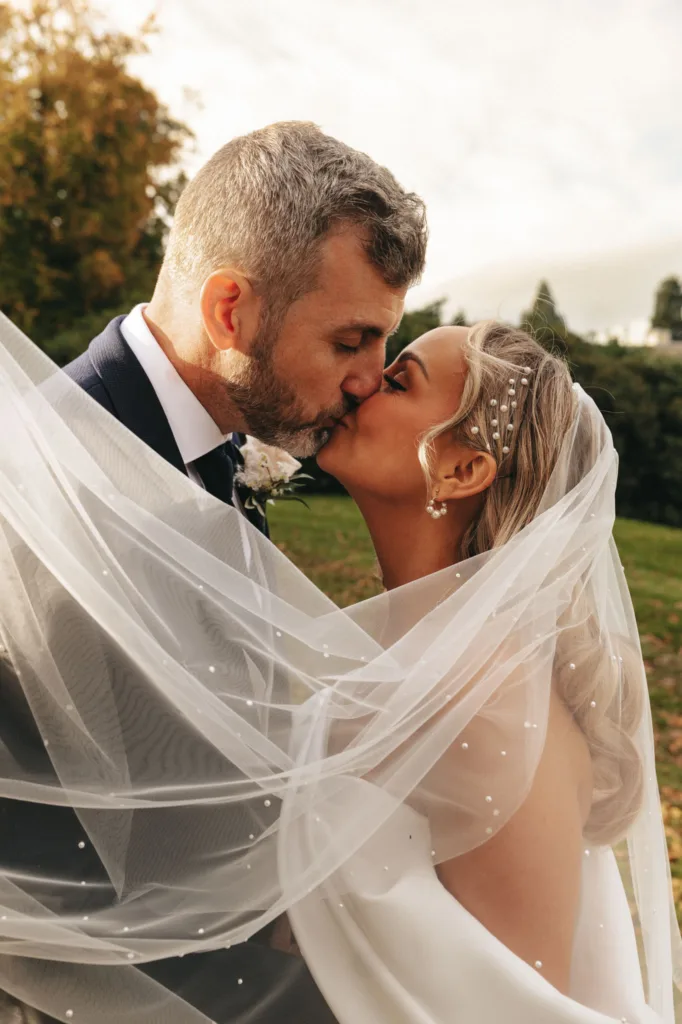 Under the radiant sun at Rudding Park, the couple shares a kiss. The bride, in her pearl-studded veil and white gown, elegantly entwines with her groom in a dark suit and boutonniere. Their embrace is framed by Harrogate's vibrant autumn foliage in the background. © Aimee Lince Photography - Wedding photographer in Lincolnshire, Yorkshire & Nottinghamshire