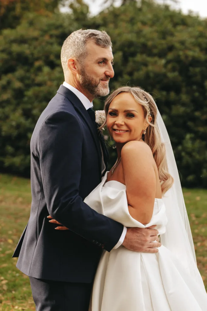 In the serene setting of Rudding Park, Harrogate, a bride and groom embrace on a grassy area with trees framing the scene. The groom, in a dark suit and beard, holds his beaming bride in an off-the-shoulder dress and veil. Both seem content amidst the natural beauty of North Yorkshire. © Aimee Lince Photography - Wedding photographer in Lincolnshire, Yorkshire & Nottinghamshire