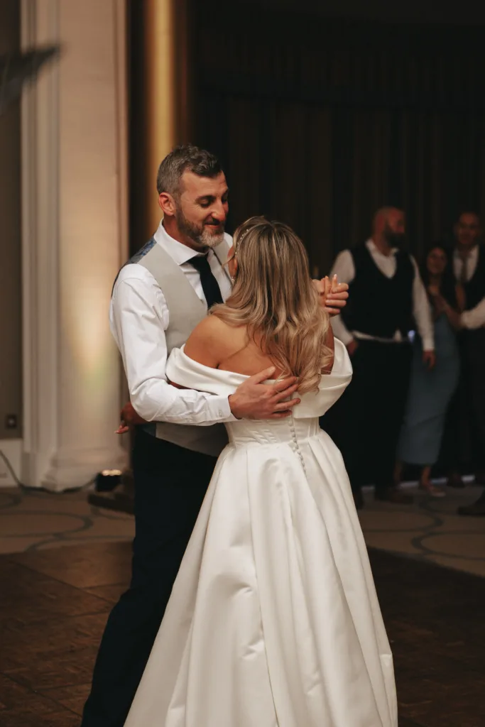 At a charming wedding in North Yorkshire, a couple dances closely. The groom, in a white shirt and gray vest, smiles warmly at the bride in her off-shoulder gown. On the wooden floor, guests in formal attire mingle, their joyful presence captured amidst a blur, including one bearded gentleman. © Aimee Lince Photography - Wedding photographer in Lincolnshire, Yorkshire & Nottinghamshire