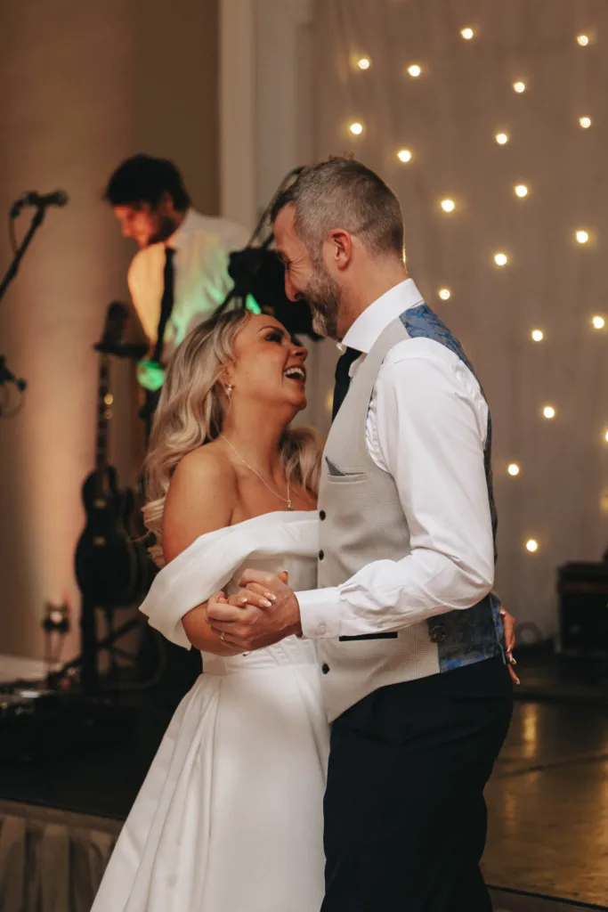 At a charming wedding reception in Harrogate, the bride and groom share a joyful dance. The bride dazzles in an off-shoulder white gown, while the groom sports a stylish blue and beige vest. Smiling at each other, they sway to guitar music under the fairy lights adorning Rudding Park's elegant walls. © Aimee Lince Photography - Wedding photographer in Lincolnshire, Yorkshire & Nottinghamshire