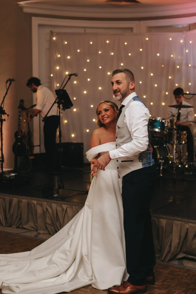At Rudding Park in North Yorkshire, a couple shares a wedding dance; the bride wears an off-shoulder white gown, while the groom sports a white shirt with a blue tie and vest. A live band with guitarist and drummer performs on a dimly lit stage adorned with string lights in the background. © Aimee Lince Photography - Wedding photographer in Lincolnshire, Yorkshire & Nottinghamshire