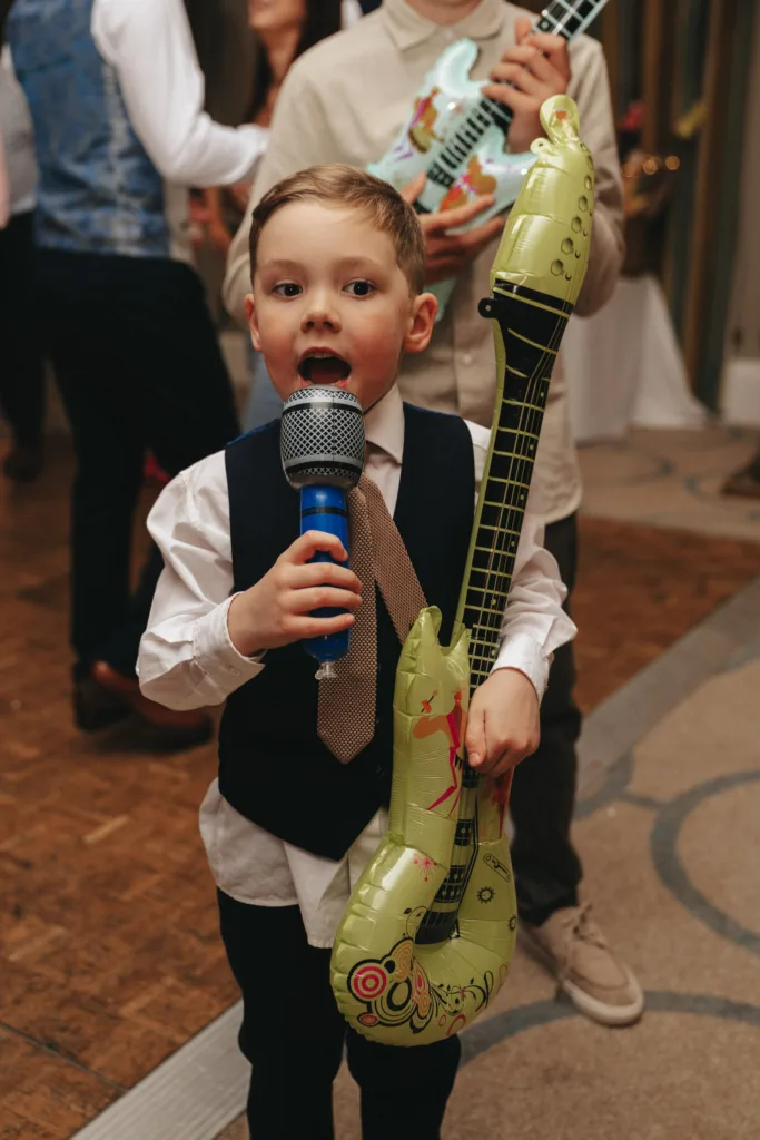 A young boy in a white shirt, dark vest, and brown tie holds a microphone and a green inflatable guitar, enthusiastically performing indoors. An adult in the background wields another inflatable guitar. The lively scene captures the spirit of a party or event in Harrogate, North Yorkshire. © Aimee Lince Photography - Wedding photographer in Lincolnshire, Yorkshire & Nottinghamshire