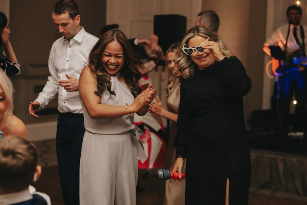 Two women are smiling and standing in a lively room at Rudding Park. One claps while the other, sporting heart-shaped sunglasses, holds a microphone. People dance in the background as a band performs on stage, capturing the festive spirit of a Harrogate celebration in Yorkshire. © Aimee Lince Photography - Wedding photographer in Lincolnshire, Yorkshire & Nottinghamshire