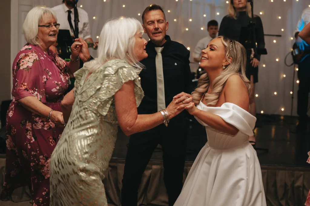 A joyful scene at a wedding reception in Harrogate shows a bride in a white gown dancing and holding hands with an elderly woman in a green dress. They are surrounded by guests, including a man in a black shirt and two women. Fairy lights adorn the backdrop at Rudding Park, enhancing the festive atmosphere. © Aimee Lince Photography - Wedding photographer in Lincolnshire, Yorkshire & Nottinghamshire