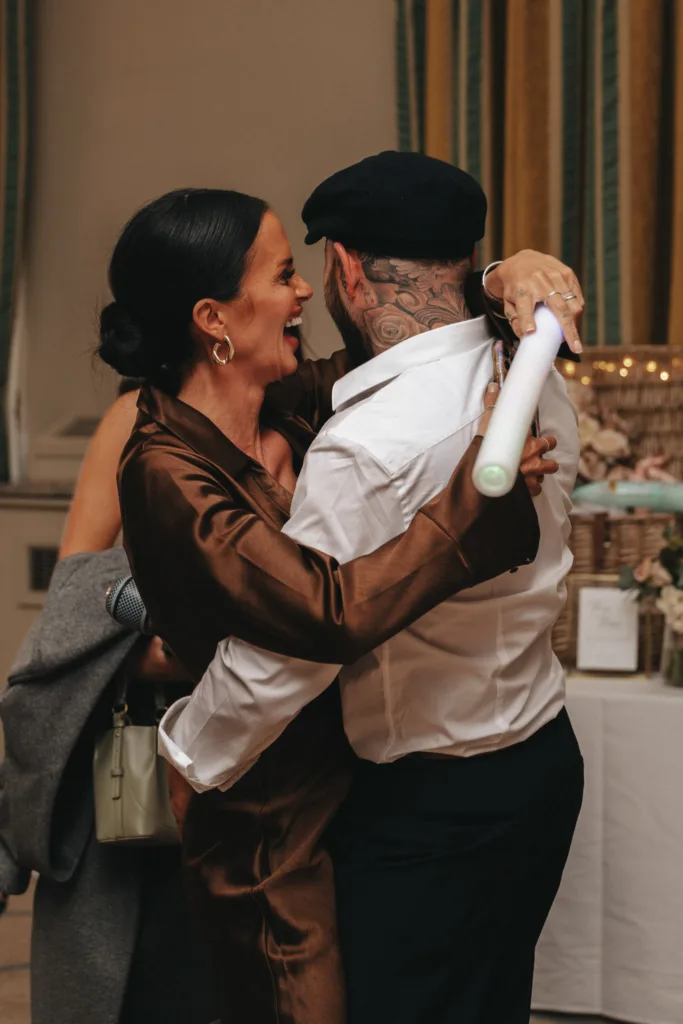 A woman in a silky brown dress and gold hoop earrings joyfully embraces a tattooed man wearing a white shirt and black cap at Rudding Park, Yorkshire. They are indoors, surrounded by a festive ambiance, with decorations and flowers on the table. The man holds a glowing stick. © Aimee Lince Photography - Wedding photographer in Lincolnshire, Yorkshire & Nottinghamshire