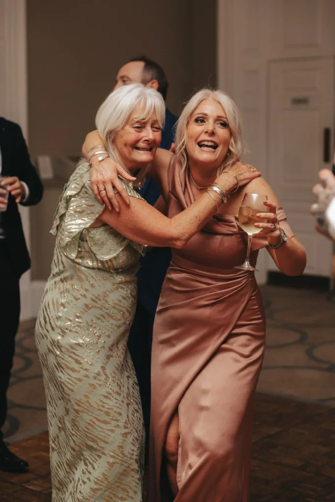 Two women joyfully pose together indoors in Harrogate, North Yorkshire. The older woman wears a light green patterned dress while the younger is in a sleeveless satin pink dress with a slit, holding a glass of wine. They smile, arms around each other, celebrating in the warmly lit room. © Aimee Lince Photography - Wedding photographer in Lincolnshire, Yorkshire & Nottinghamshire