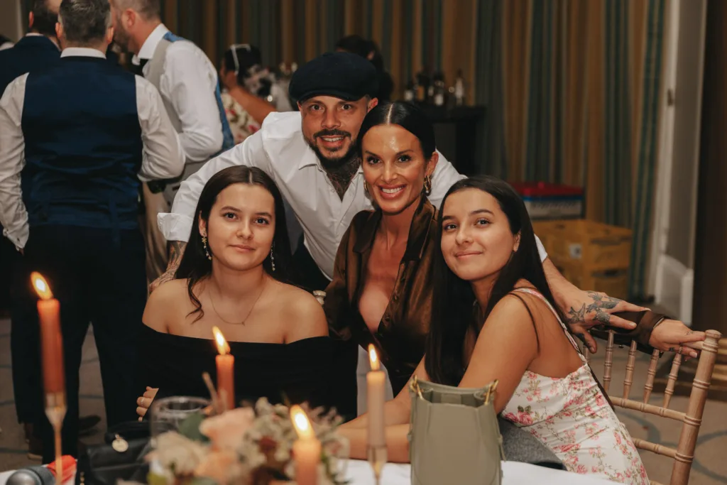 A group of four smiling adults is gathered around a table at an event in Harrogate. The table has lit candles and a floral centerpiece. Behind them are other guests, some with their backs turned. The setting appears festive, with warm lighting and curtains in the background, reminiscent of Rudding Park's charm. © Aimee Lince Photography - Wedding photographer in Lincolnshire, Yorkshire & Nottinghamshire
