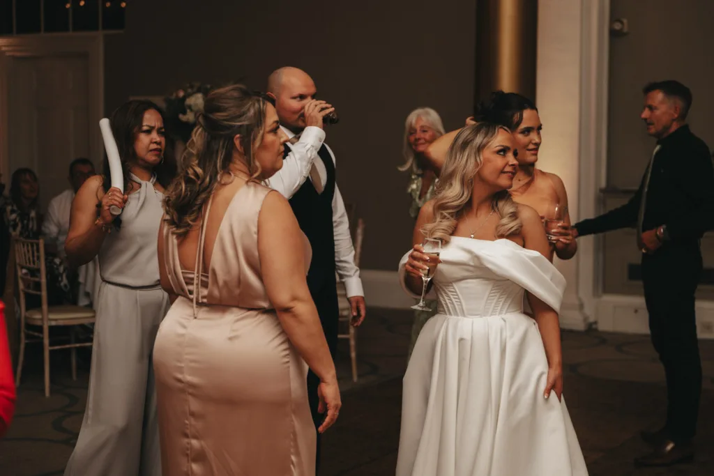 A bride in an off-shoulder white gown stands smiling with a drink in her hand, surrounded by guests in elegant attire, including a man in a vest drinking from a glass and women in satin dresses. The scene unfolds indoors with soft lighting and warm decor, reminiscent of a Harrogate celebration. © Aimee Lince Photography - Wedding photographer in Lincolnshire, Yorkshire & Nottinghamshire