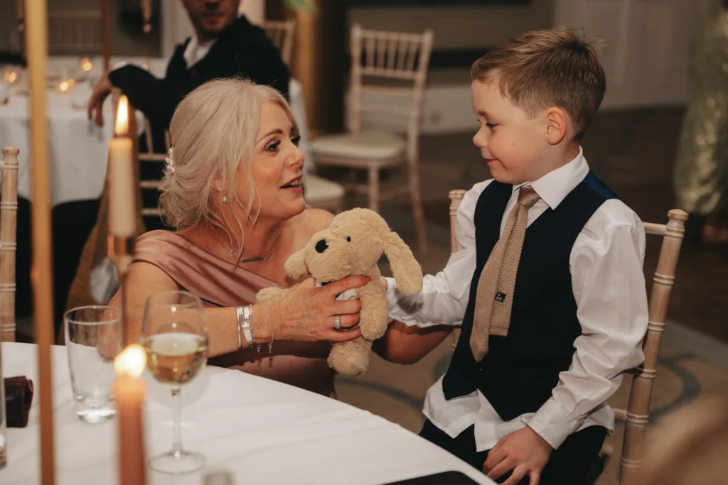 An elderly woman with gray hair holds a stuffed bunny and smiles at a young boy in a suit with a tie. They sit at a table adorned with candles and glasses of wine and water, capturing the essence of an elegant celebration in Rudding Park, North Yorkshire. © Aimee Lince Photography - Wedding photographer in Lincolnshire, Yorkshire & Nottinghamshire