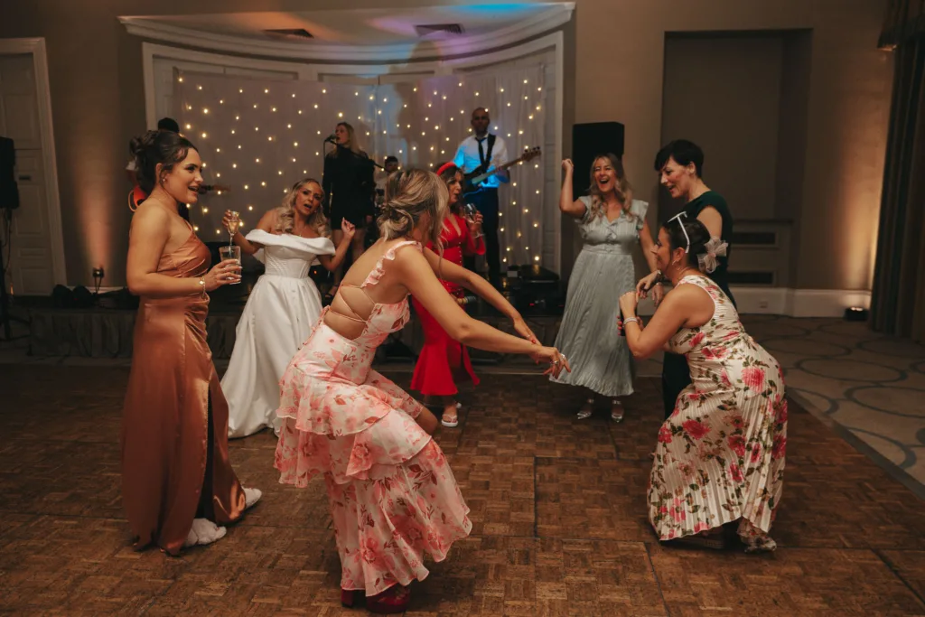 A group of women in vibrant dresses dance joyfully at a party in Rudding Park. Behind them, a live band plays against a backdrop of twinkling lights. The wooden floor and lively atmosphere capture the festive spirit, with guests laughing and moving energetically to the music in Yorkshire's enchanting setting. © Aimee Lince Photography - Wedding photographer in Lincolnshire, Yorkshire & Nottinghamshire