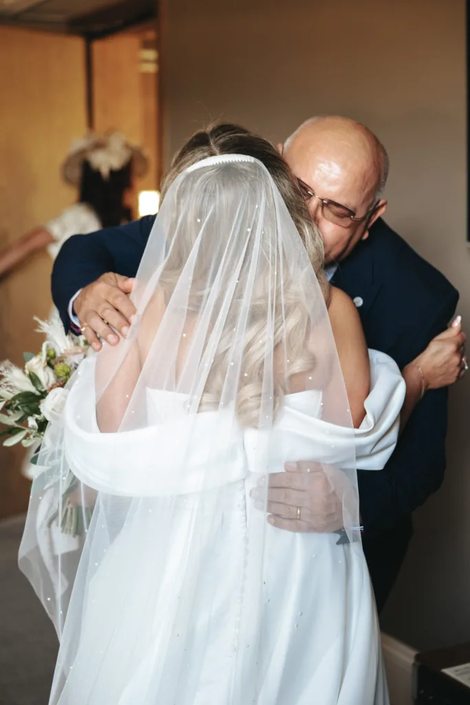 A bride in a white gown and veil hugs an older man wearing glasses and a dark suit at Rudding Park, North Yorkshire. He appears emotional as she holds a bouquet of white flowers. A woman in a hat is blurred in the background near a wooden door, suggesting an intimate indoor setting. © Aimee Lince Photography - Wedding photographer in Lincolnshire, Yorkshire & Nottinghamshire
