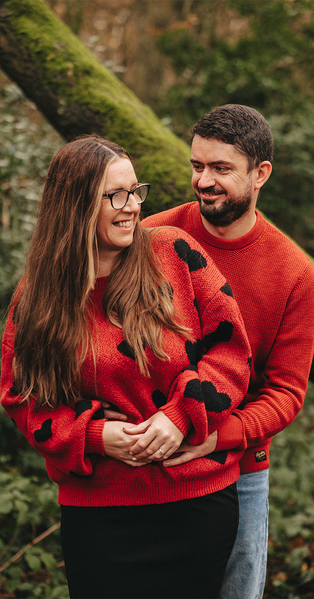 A smiling couple stands in a forest at Winter Weelsby Woods. Rebecca, with long brown hair and glasses, wears a red sweater with a black pattern. Daniel, with short dark hair and a beard, wears a plain red sweater and holds her from behind. Both appear happy during their engagement shoot, surrounded by greenery and a mossy log. © Aimee Lince Photography