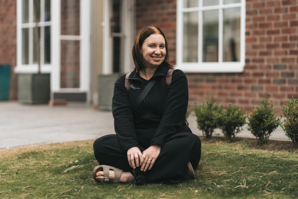 Aimee Lince Photography A woman with shoulder-length auburn hair smiles while sitting cross-legged on a grassy patch, her photographer's eye catching every detail. She wears a black sweater, pants, and brown sandals. Behind her, there's a brick building with a large window and door, surrounded by small shrubs. © Aimee Lince Photography - Wedding photographer in Lincolnshire, Yorkshire & Nottinghamshire
