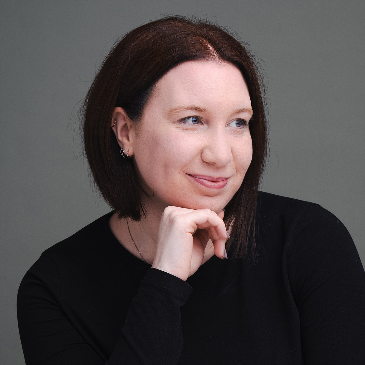 A female wedding photographer with short, dark brown hair is beautifully captured in a black top and small hoop earrings. They're resting their chin on their hand, gazing to the side with a slight smile. The plain grey background adds a calm and contemplative feel to this engagement photography moment. © Aimee Lince Photography