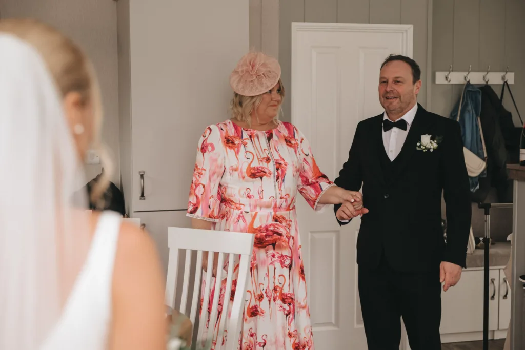 Brackenborough Hotel Wedding Photographer - Mollie & Ryan © Aimee Lince Photography (217) A woman in a pink flamingo-patterned dress and pink fascinator stands next to a man in a black tuxedo with a bow tie, holding hands. Amidst the celebration, it’s a beautiful moment where mother-daughter memories enhance the wedding day. A white dress is seen blurred in the foreground. © Aimee Lince Photography - Wedding photographer in Lincolnshire, Yorkshire & Nottinghamshire