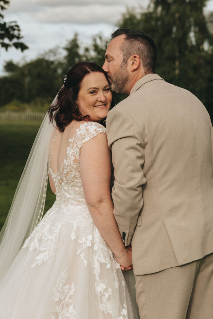 A bride and groom stand outdoors, smiling and holding hands. The bride wears a white lace wedding dress with a veil, and the groom wears a light beige suit. He gently kisses her on the forehead. They are surrounded by lush greenery and a white fence is visible in the background. © Aimee Lince Photography
