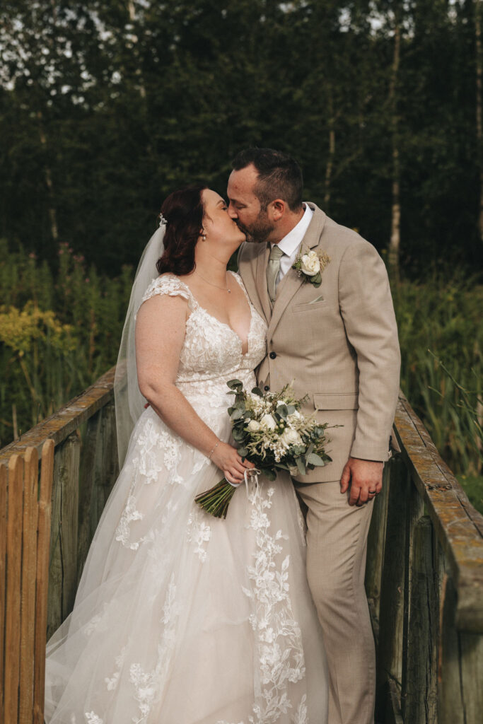 Bride in a lace wedding dress and groom in a beige suit share a kiss while standing on a wooden bridge. The bride holds a bouquet of white and green flowers. Blurred greenery and trees are in the background, creating a serene, natural setting. © Aimee Lince Photography
