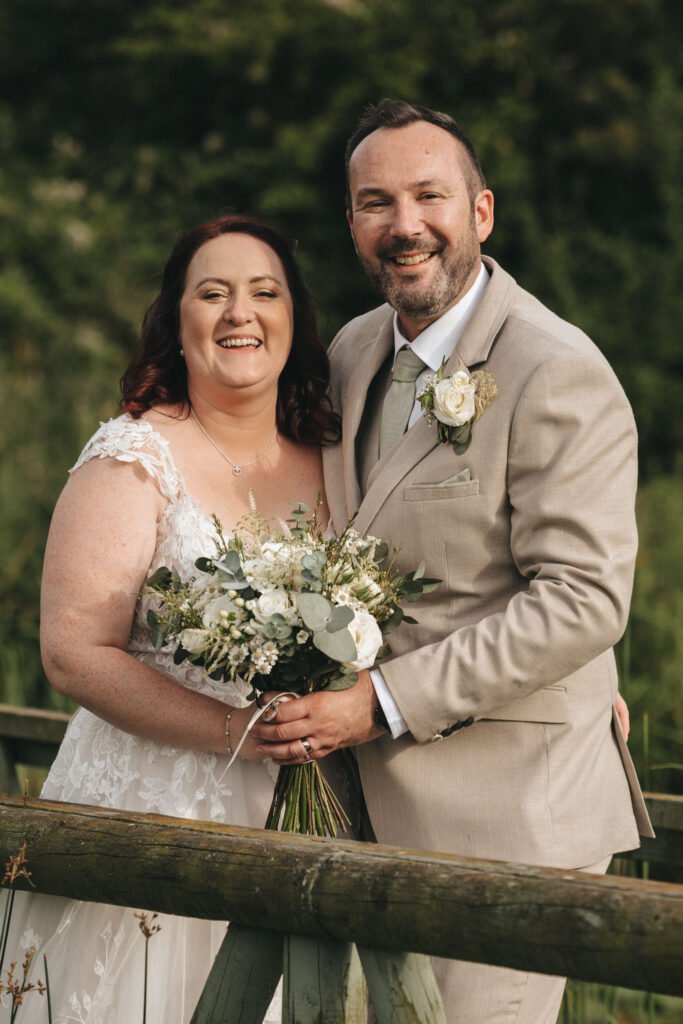 A couple in wedding attire stands outdoors, smiling. The bride holds a bouquet with white flowers and greenery, wearing a lace gown. The groom is in a beige suit with a boutonniere. They lean on a wooden fence, set against a backdrop of lush greenery, suggesting a garden setting. © Aimee Lince Photography