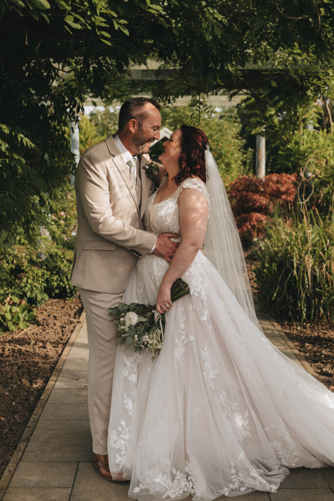 A couple stands on a garden pathway under a leafy canopy. The groom, in a beige suit, embraces the bride, who wears a flowing white gown with lace details and a veil. She holds a bouquet of white flowers. They look lovingly at each other, surrounded by lush greenery and sunlight. © Aimee Lince Photography
