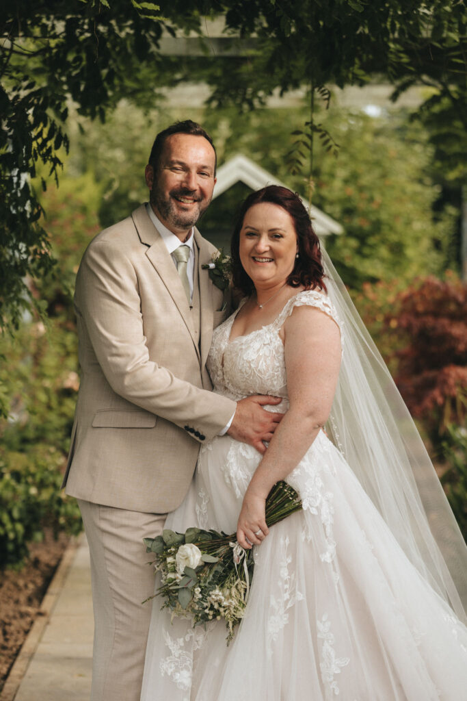 A happy couple poses outdoors on their wedding day. The groom is in a beige suit and white shirt. The bride is wearing a lace gown with a veil and holding a bouquet. They stand closely under green foliage, smiling. The background features lush greenery and a small building. © Aimee Lince Photography