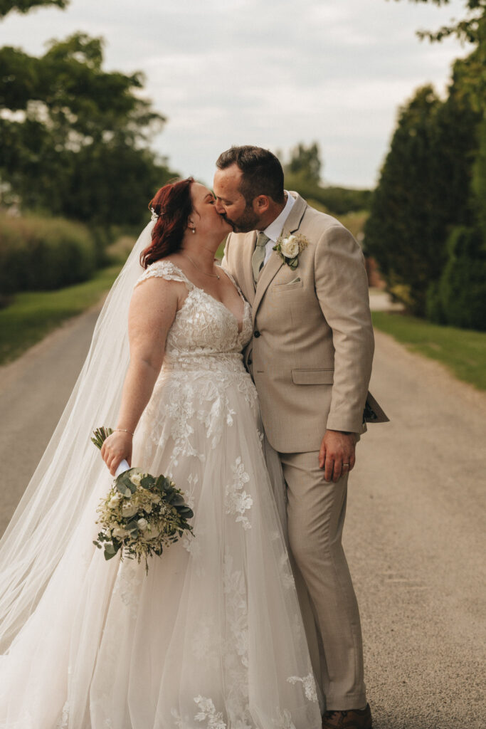 A bride and groom kiss on a rural road, surrounded by greenery. The bride wears a white lace gown and veil, holding a bouquet, while the groom is in a beige suit with a floral boutonniere. The scene captures a romantic moment with trees and cloudy sky in the background. © Aimee Lince Photography