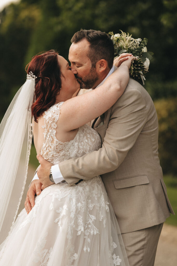 A bride and groom share a kiss outdoors. The bride wears a white lace gown and veil, holding a bouquet. The groom is in a light beige suit and white shirt. They embrace closely, with greenery and blurred foliage in the background, capturing a romantic and intimate moment. © Aimee Lince Photography
