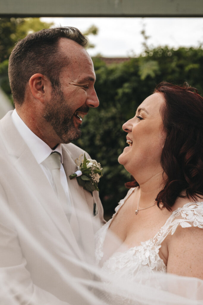 A bride and groom laugh joyfully as they look into each other's eyes. The groom is wearing a light beige suit with a boutonniere, and the bride is in a white lace dress. They are standing close together, with greenery in the background. The moment is captured in a candid and happy atmosphere. © Aimee Lince Photography