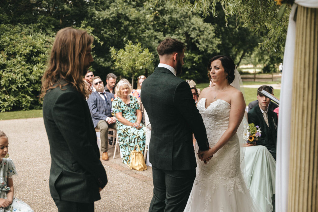 A bride and groom stand facing each other, holding hands, during an outdoor wedding ceremony. The groom wears a dark suit and the bride wears a strapless lace gown. Guests seated in the background watch the couple. Greenery surrounds the area, creating a natural setting. © Aimee Lince Photography at The Dower House Hotel