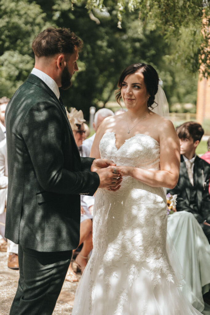 A bride and groom stand facing each other during an outdoor wedding ceremony. The bride is in a white lace gown with a veil, while the groom is in a dark suit. They hold hands, surrounded by seated guests. Sunlight filters through tree leaves, creating dappled shadows on their clothes. © Aimee Lince Photography at The Dower House Hotel