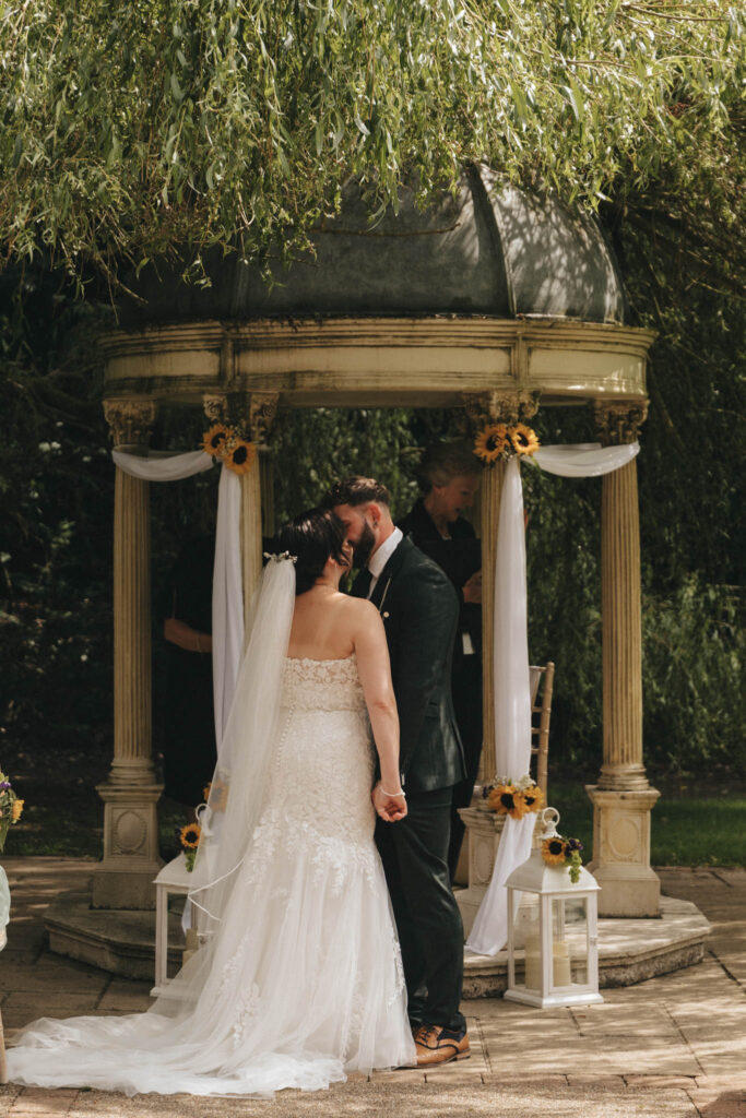 A couple shares a kiss during their wedding ceremony under a decorated gazebo. The bride wears a strapless white gown and veil, while the groom is in a dark suit. The gazebo is adorned with white fabric and sunflowers. A person stands in the background, partially visible behind the couple. © Aimee Lince Photography at The Dower House Hotel