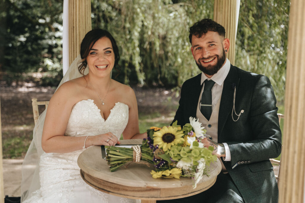 A bride and groom sit at a small round table under a canopy of greenery. The bride, in a strapless lace gown, and the groom, in a dark suit with a polka-dot tie, both smile at the camera. A bouquet with sunflowers and greenery is on the table. The background is lush with foliage. © Aimee Lince Photography at The Dower House Hotel