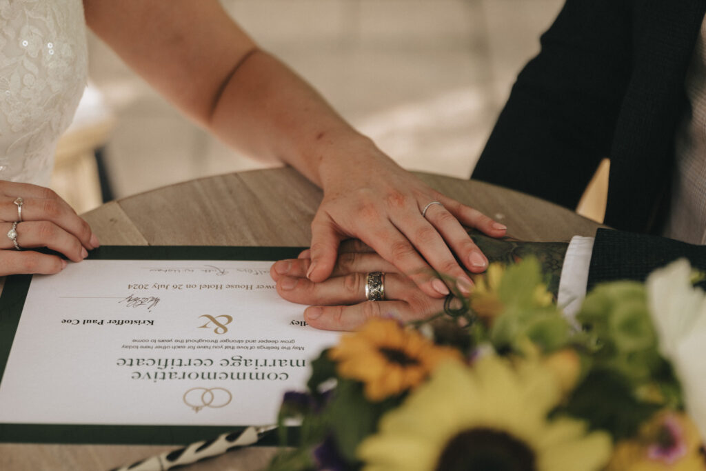 Close-up of a couple's hands resting on a table with a marriage certificate. The woman's hand, with a wedding ring, gently holds the man's hand, which also wears a ring. A bouquet with sunflowers is partially visible in the foreground. The certificate reads "Commonwealth Marriage Certificate. © Aimee Lince Photography at The Dower House Hotel