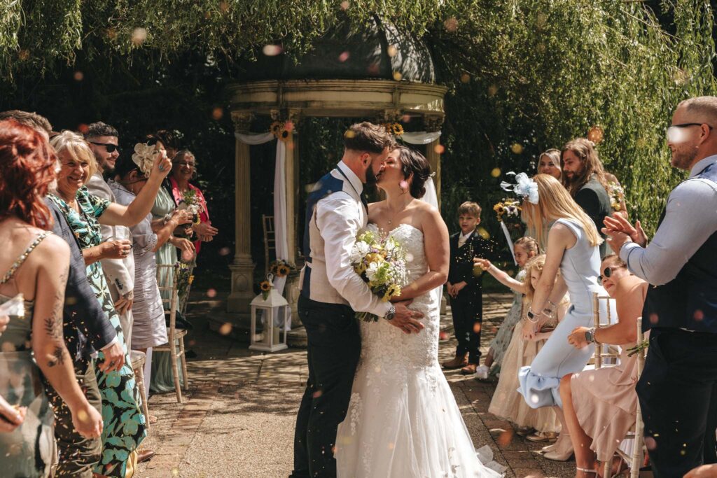 A bride and groom share a kiss outdoors surrounded by smiling guests. The bride wears a white gown and holds a bouquet, while the groom is in a white shirt with a blue vest. Guests cheer, some throwing confetti. Sunlight filters through trees, creating a festive, joyful atmosphere. © Aimee Lince Photography at The Dower House Hotel