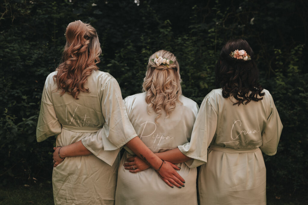 Three women with long hair, wearing matching light green robes, stand arm in arm facing away from the camera. Each robe has a name written on the back. They are outdoors, in front of dark green foliage, each with flower-adorned hairstyles. © Aimee Lince Photography at The Dower House Hotel