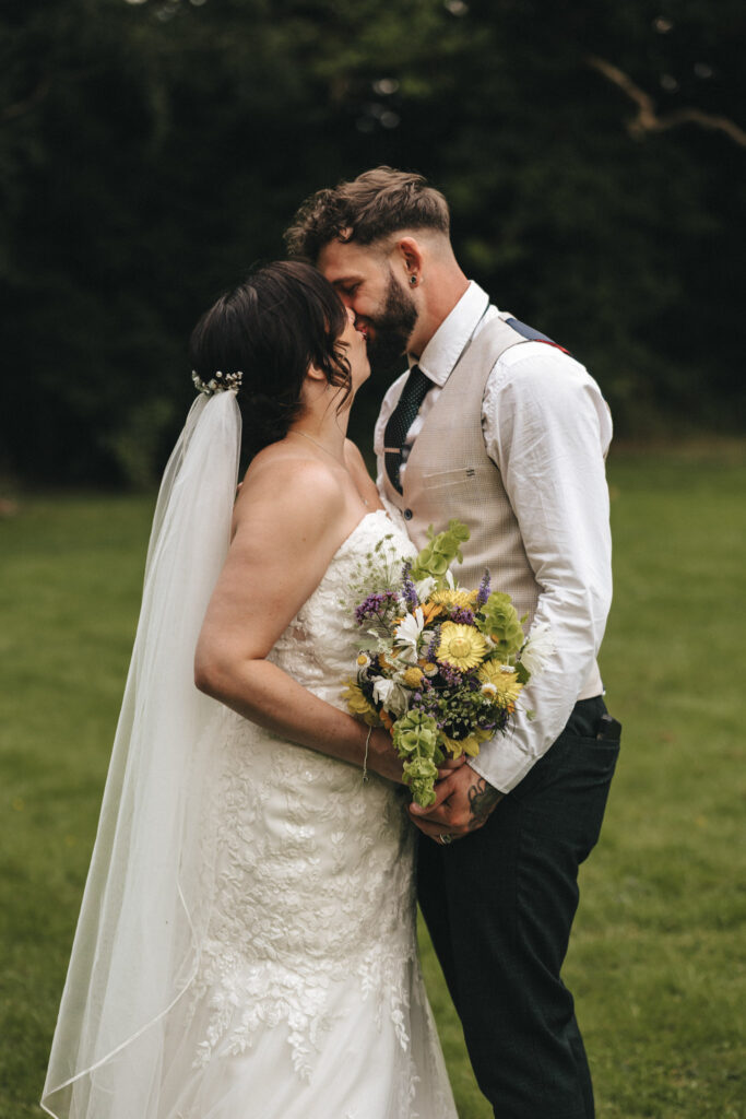 A bride and groom share a tender moment outdoors. The bride is in a lace wedding dress with a veil, holding a vibrant bouquet of yellow and purple flowers. The groom is in a white shirt with suspenders and a tie. They stand closely together on grass with trees in the background. © Aimee Lince Photography at The Dower House Hotel