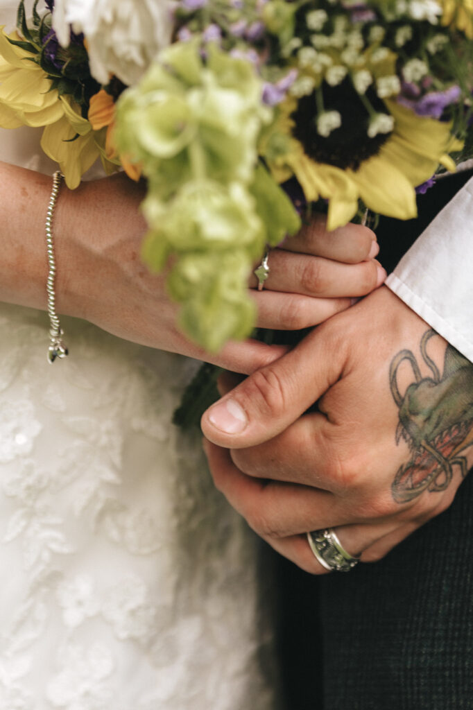 Close-up of a bride and groom holding hands. The bride, in a lace gown, wears a charm bracelet. The groom has a tattoo of a dragon on his hand and wears a silver ring. They are holding a bouquet with sunflowers, white roses, and greenery, symbolizing their connection and joy. © Aimee Lince Photography at The Dower House Hotel