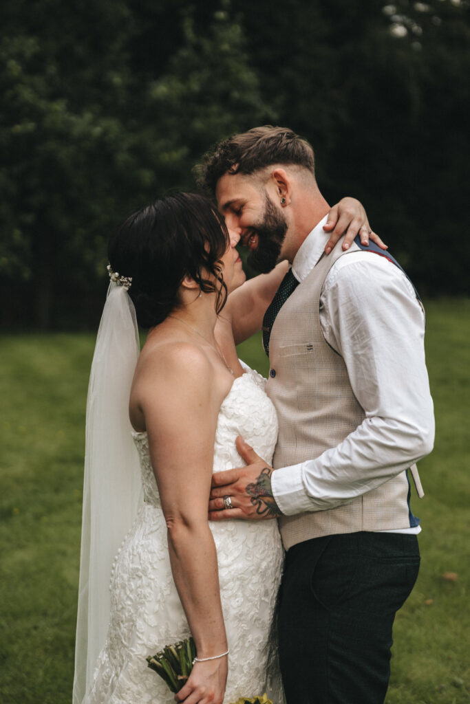 A couple embraces outdoors on a lush green lawn. The bride, in a white lace wedding dress and veil, holds a bouquet. The groom wears a beige vest over a white shirt and black pants. They touch foreheads affectionately, both smiling with eyes closed. Trees are in the background. © Aimee Lince Photography at The Dower House Hotel