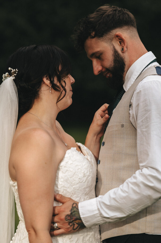 A bride and groom stand closely, gazing at each other lovingly. The bride wears a white lace strapless gown and a veil with floral accents. The groom, with a trimmed beard, wears a beige vest over a white shirt and holds the bride's waist. They are outdoors with greenery in the background. © Aimee Lince Photography at The Dower House Hotel