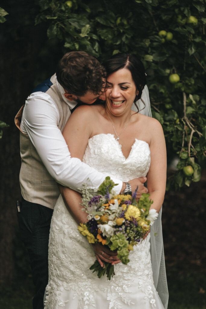 A joyful bride in a white lace strapless gown holds a colorful bouquet of flowers. She's being hugged from behind by a man in a white shirt and beige vest. They are surrounded by lush greenery, and both are smiling. The scene conveys a sense of love and happiness. © Aimee Lince Photography at The Dower House Hotel