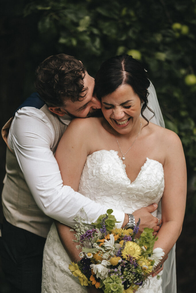 A bride in a white lace dress and veil holds a colorful bouquet, smiling joyfully as a groom in a white shirt and vest embraces her from behind. They are surrounded by green foliage, with the groom playfully nuzzling her neck. The scene conveys happiness and intimacy. © Aimee Lince Photography at The Dower House Hotel